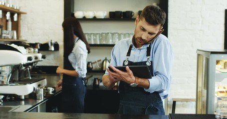 Caucasian attractive waiter speaking on the telephone and using tablet computer at the bar, brunette waitress making coffee behind. - Powered by Adobe