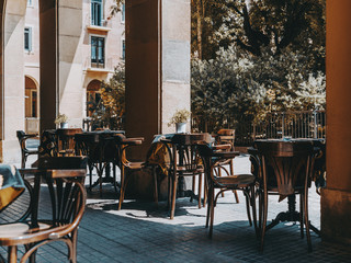 Cozy outdoor cafe with empty wooden tables covered with a tablecloth with metal flower pots and ashtries on it, chairs near; exterior of an empty street bar on the daytime waiting for its visitors