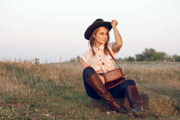 Girl cowgirl in jeans boots shirt and with a bag in a box with a wheel by a tree