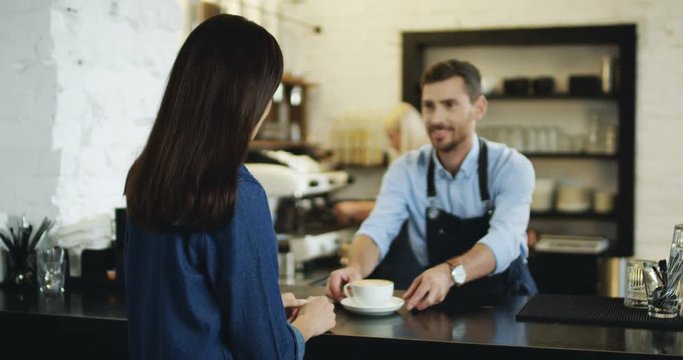 Handsome Caucasian barmen serving coffee to the pretty brunette woman at the bar, then she turning and smiling to the camera.