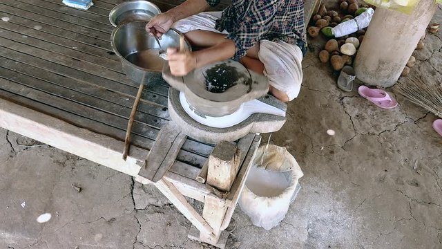 upper view on woman sat cross-legged using hand-turned millstone to grind wet rice for making soaked rice flour