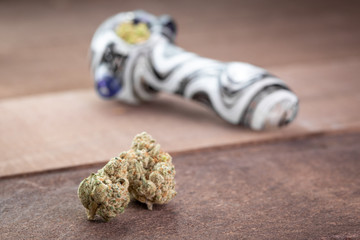 Marijuana buds on wood, with a packed cannabis smoking pipe in the background