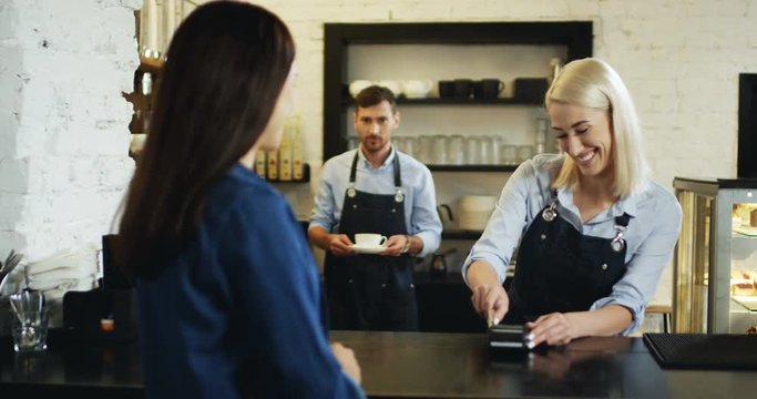 Rear of the brunette woman paying with a credit card to the blonde pretty waitress at the bar, while waiter bringing her coffee.