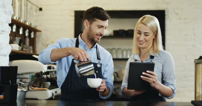 Beautiful blonde woman waitress watching video on the tablet device while attractive waiter pouring milk in the cup of coffee at the bar.