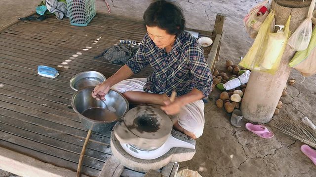 Woman - sat cross-legged on a bamboo sitting platform - using hand-turned millstone to grind wet rice for making soaked rice flour and taking the millstone away (upper view)