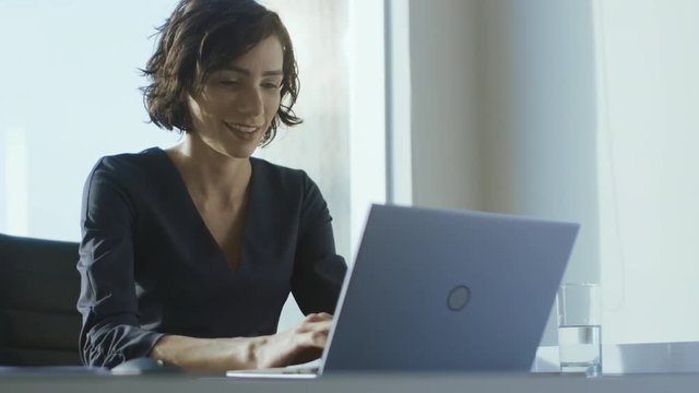 Confident Female Executive Works on a Laptop Sitting at Her Desk in Modern Office with Big City View. Smiling Successful Businesswoman Uses Laptop.