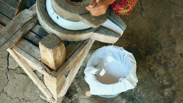 upper view on woman sat cross-legged using hand-turned millstone to grind wet rice for making soaked rice flour