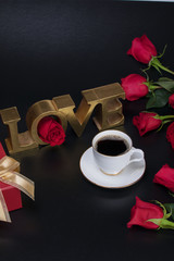 Red gift box with gold ribbon, red roses, word love and white cup of coffee on black background