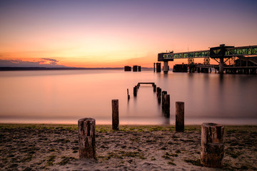 Fototapeta premium Long exposure of pilings leading in to a calm sea at sunset, with a ferry terminal in background