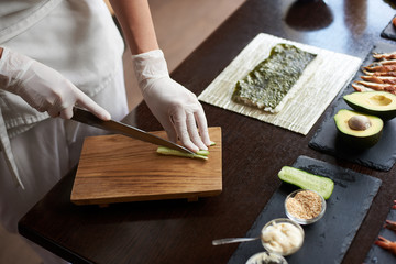 Close-up view of process of preparing delicious rolling sushi in restaurant. Female hands in disposable gloves slicing cucumber on wooden board.