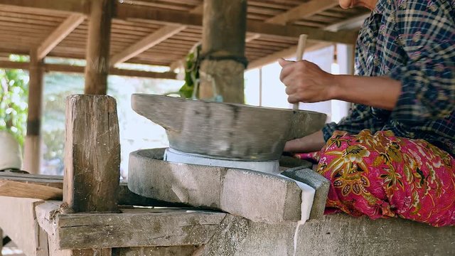 woman sat cross-legged using hand-turned millstone to grind wet rice for making soaked rice flour