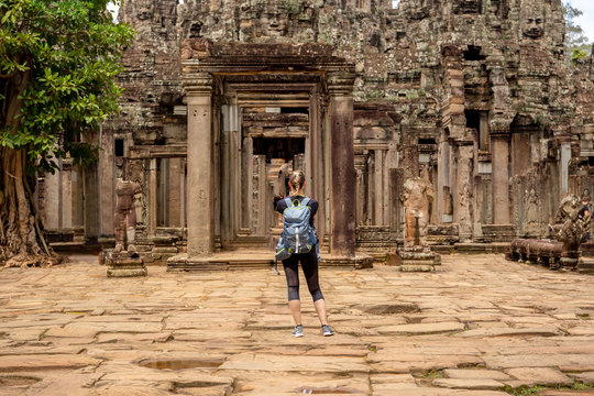 Young female tourist with backpack taking picture of ancient Bayon temple in Siem Reap Cambodia with cell phone.
