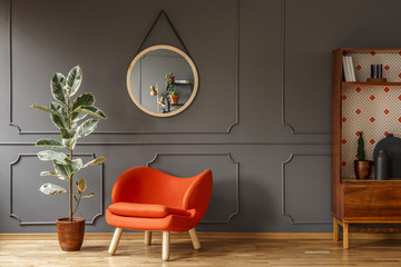 Bright orange armchair, a retro wooden cabinet and a mirror on a gray wall with molding in a...