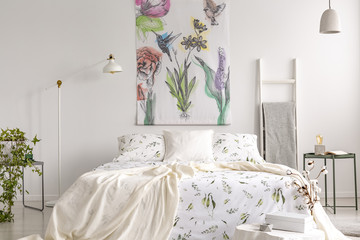 Wall art of flowers and birds painted on a fabric above a bed which is dressed in green plants pattern on white bedding in a fresh looking bedroom interior. Real photo.