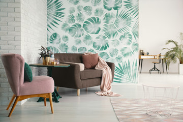 Pink blanket on brown couch next to table and armchair in living room interior with leaves. Real...