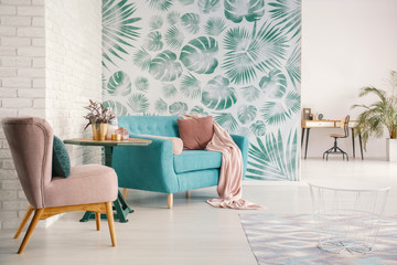 Flowers on table between pink armchair and turquoise sofa in flat interior with green wallpaper....