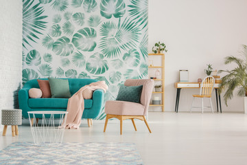 Pink chair next to blue couch against green wallpaper in spacious flat interior with stool. Real...