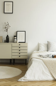 A monochromatic beige and white bedroom interior with a view at a bed and a drawer cabinet standing on a wooden floor. Real photo.