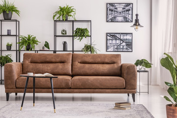 Table next to leather couch in natural white living room interior with posters and plants. Real photo