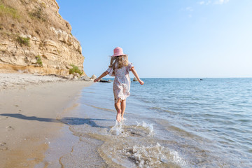 Child girl in summer dress with straw hat is running barefoot in shallow sea water