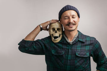 Stylish happy man with mustache and knitted hat, holding scull while posing indoors, with copy...