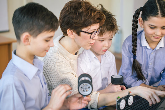 Curious clever pupils with the help of their female teacher doing a group project programming homemade robot using laptops on extracurricular classes
