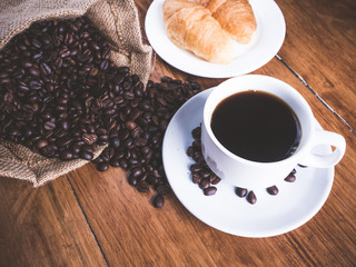 Coffee is a brewed drink prepared from roasted coffee beans, Coffee cup, coffee beans and croissant on a wooden table