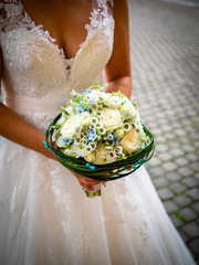 The bride holds the bouquet with white roses and little blue flowers, surrounded by green leaves and blue pearls, white lace dress, and daisies