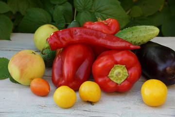 tomatoes red and yellow peppers red hot chili peppers apples cucumbers. green leaves on a white wooden table.
