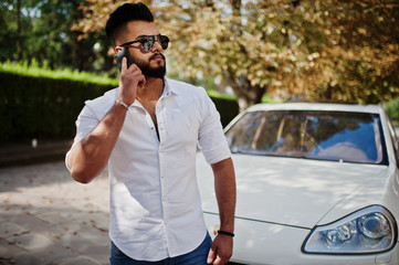 Stylish tall arabian man model in white shirt, jeans and sunglasses posed at street of city. Beard rich attractive arab guy against white suv car, speaking on mobile phone.