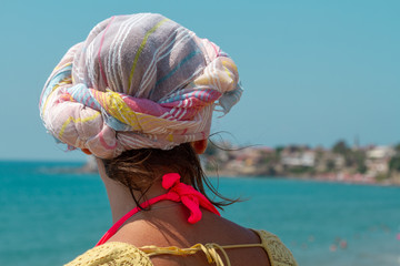 A young tanned woman in a turban on her head stares into the distance, to the blue sea, standing with her back to the photographer. A girl in a turban looks from a height to the azure sea.