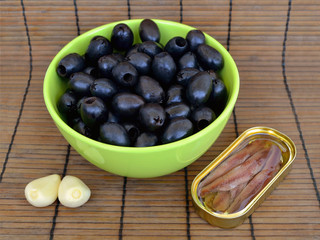 Ingredients for tapenade: stoned black olives in a green   bowl, anchovy fillets in a yellow oval tin can and two big   cloves of garlic, front view