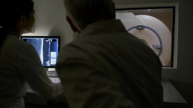 Rear view of two doctors working on computer at desk, performing MRI examination of patient and discussing procedure