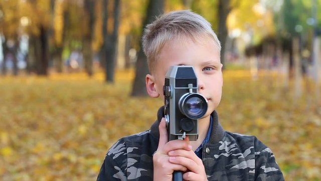 Closeup view of cute face of blond white kid holding vintage black camera for shooting video. Boy isolated on golden autumnal nature background. Real time full hd video footage.
