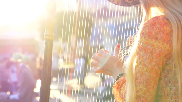 SLOW MOTION, CLOSE UP, LENS FLARE, COPY SPACE: Unrecognizable blonde haired woman playing the harp at sunset. Unknown female street artist plucking the strings of a harp and entertaining tourists.