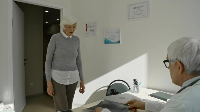 Elderly woman walking into doctor’s office and talking to senior practitioner while he examining x-ray Image