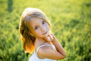 close-up portrait of beautiful little child in white dress posing in green field and looking at camera