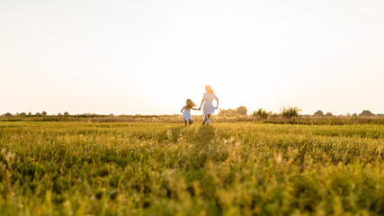 rear view of mother and daughter running in green meadow with sunset on background