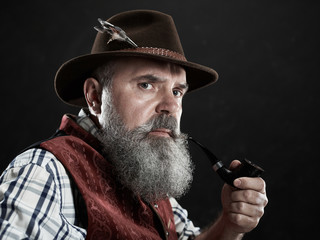 dramatic portrait of gray bearded senior man in hat smoking tobacco pipe. view of Austrian, Tyrolean, Bavarian old man in national traditional costume in retro style.