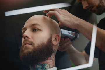 Bearded skinhead man in barbershop. Barber shaves head with electric trimmer.