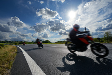 Three motion blurred motorcycles riding on an asphalt road in a rural landscape under a glowing sun and dramatic clouds