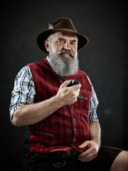 dramatic portrait of gray bearded senior man in hat smoking tobacco pipe. view of Austrian, Tyrolean, Bavarian old man in national traditional costume in retro style.