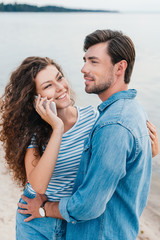 man hugging his smiling girlfriend while she talking on smartphone near sea