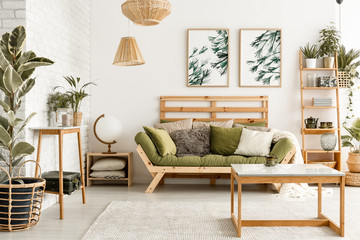 Wooden table on carpet in front of green couch in living room interior with plants and posters....