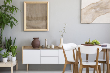 Dining table with fresh fruits and white wooden chairs in real photo of light grey room interior with two posters, green plants and small cupboard with decor