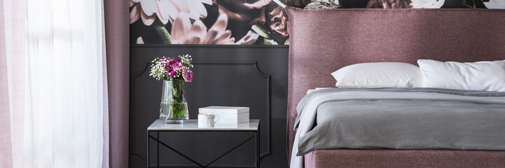 Real photo of marble bedside table with tea cup, box and fresh flowers standing by the bed in dark grey room interior with window with drapes
