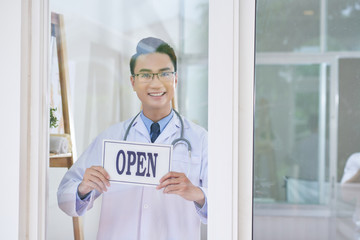 Handsome Asian guy in medical apparel smiling and looking at camera while standing behind glass in office and holding open sign