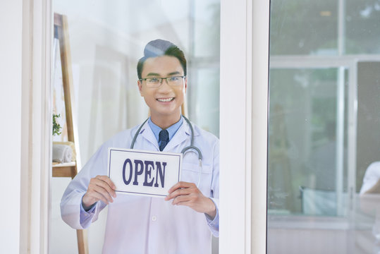 Handsome Asian guy in medical apparel smiling and looking at camera while standing behind glass in office and holding open sign