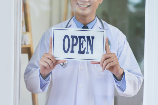 Unrecognizable male in medical apparel smiling and holding open sign while standing behind glass in office
