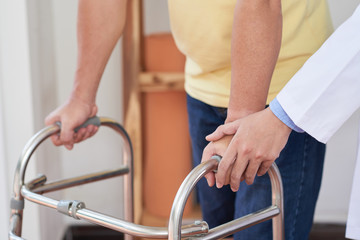 Anonymous medical practitioner assisting injured man with walking frame during rehabilitation...